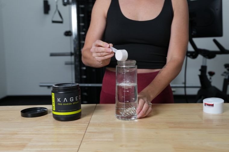 A woman pouring a scoop of Kaged Pre-Kaged Sport into a water bottle