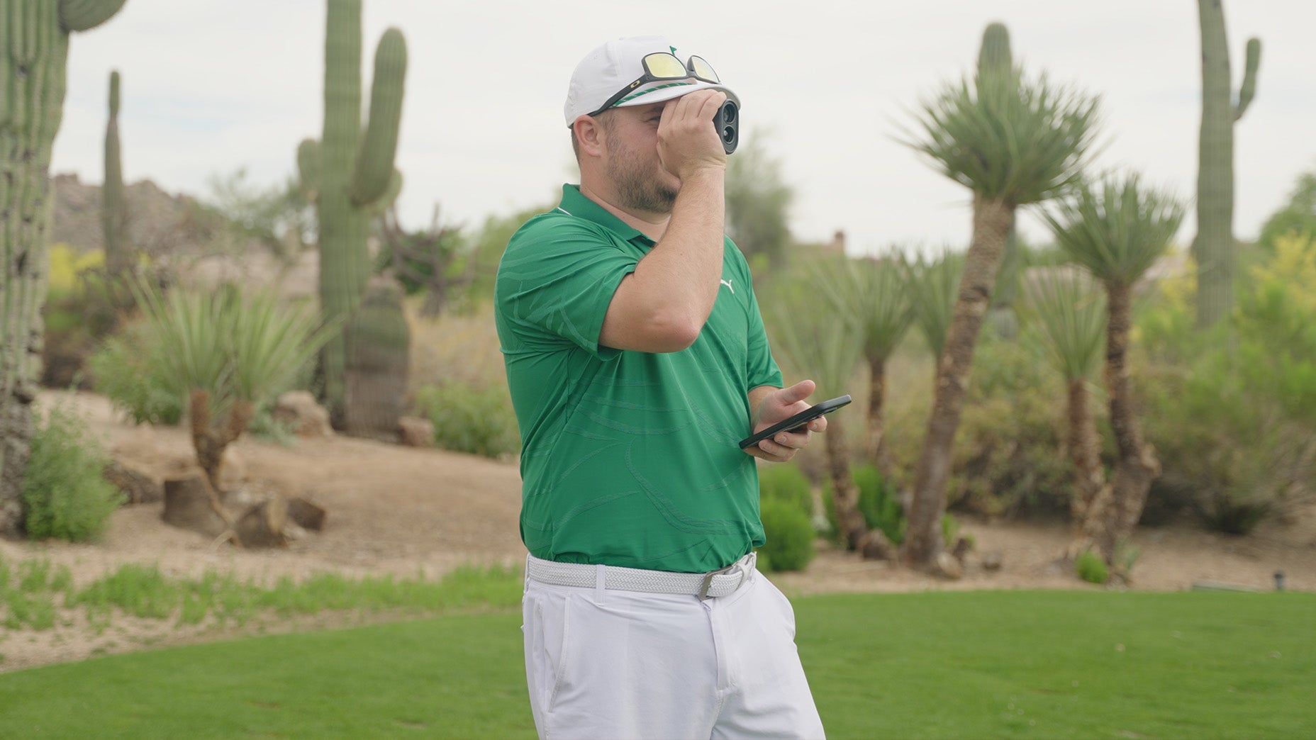 A man in a green shirt and white pants stands on a golf course with cacti in the background, looking through a rangefinder and holding a smartphone in his other hand.