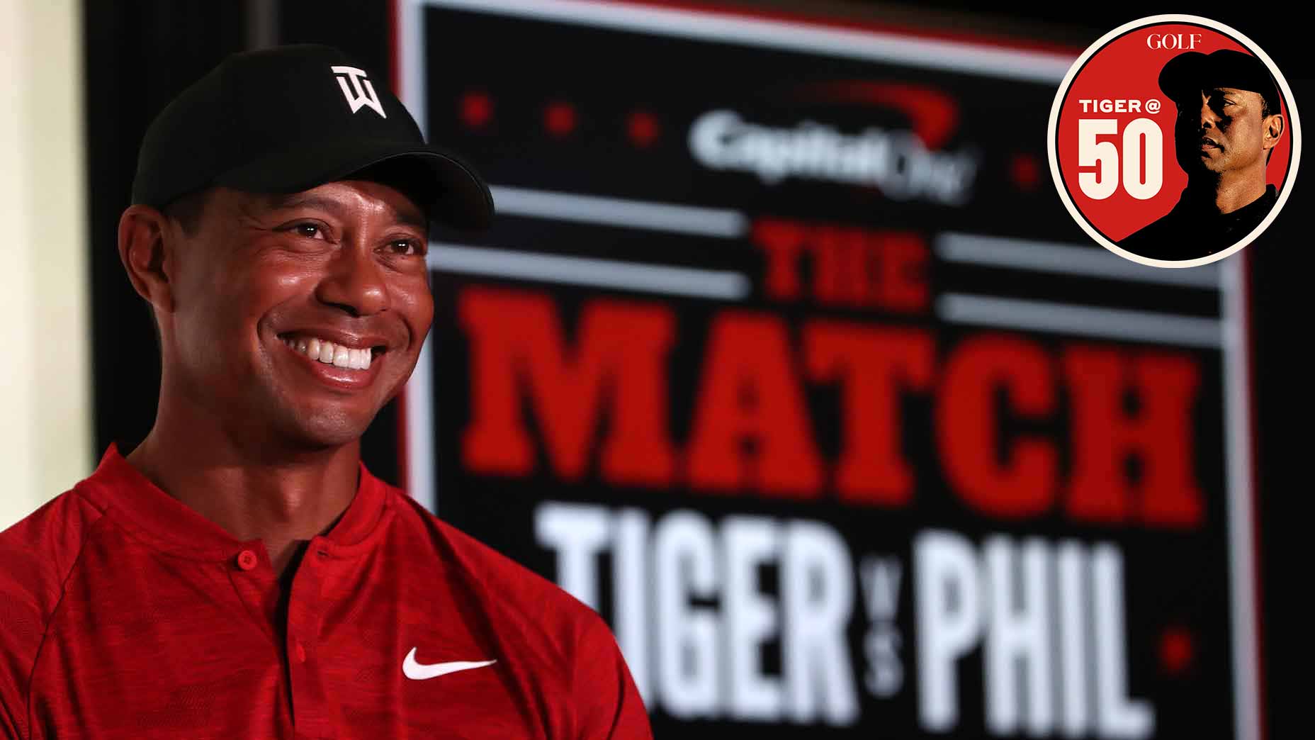 tiger woods smiles in front of the match tiger vs phil signage in red shirt and black hat.