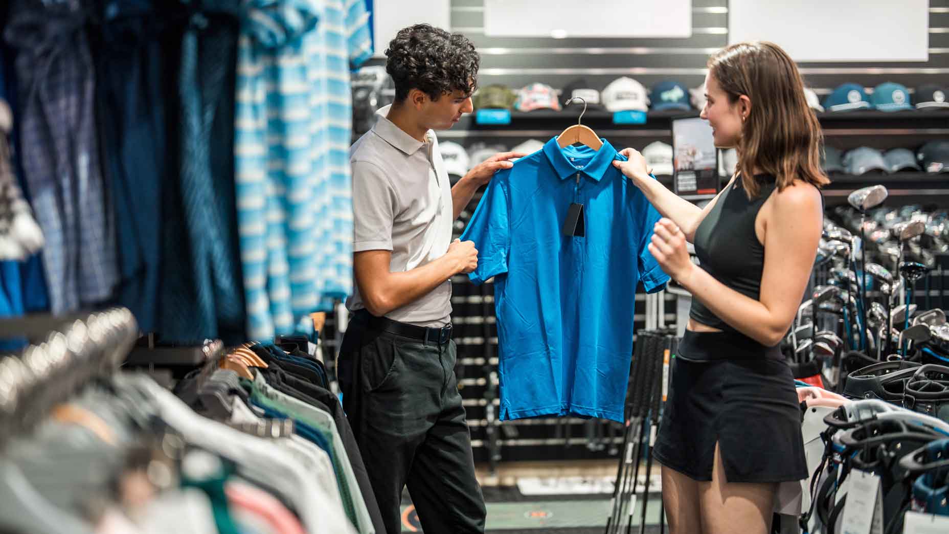 Young diverse couple, male and female, excitedly browsing golf apparel in a sports store. The man holds a blue golf shirt, while the woman observes. Both are casually dressed in summer outfits, exploring options amidst golf gear displays.